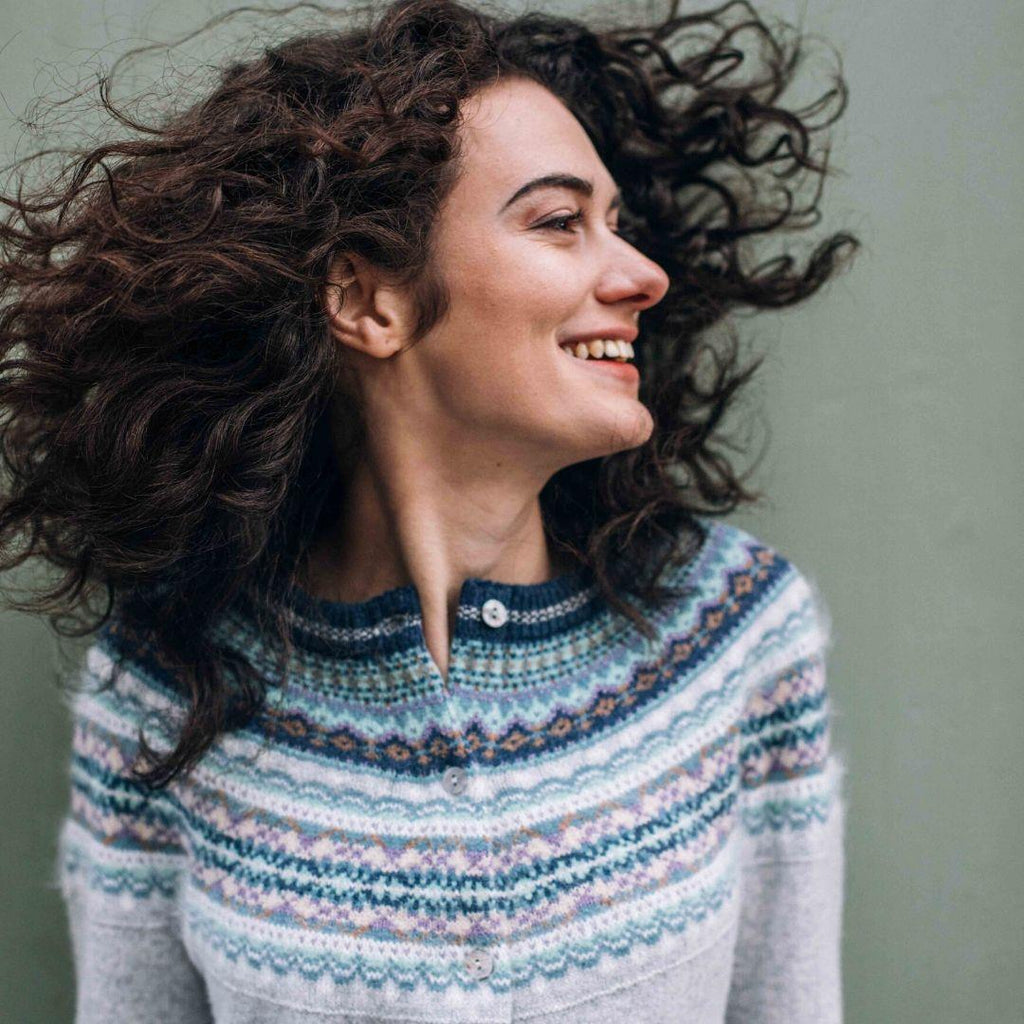 a woman smiling with her hair blowing in the wind, wearing a fairisle knit sweater in 'arctic' colours of grey, blue, white, and lavender